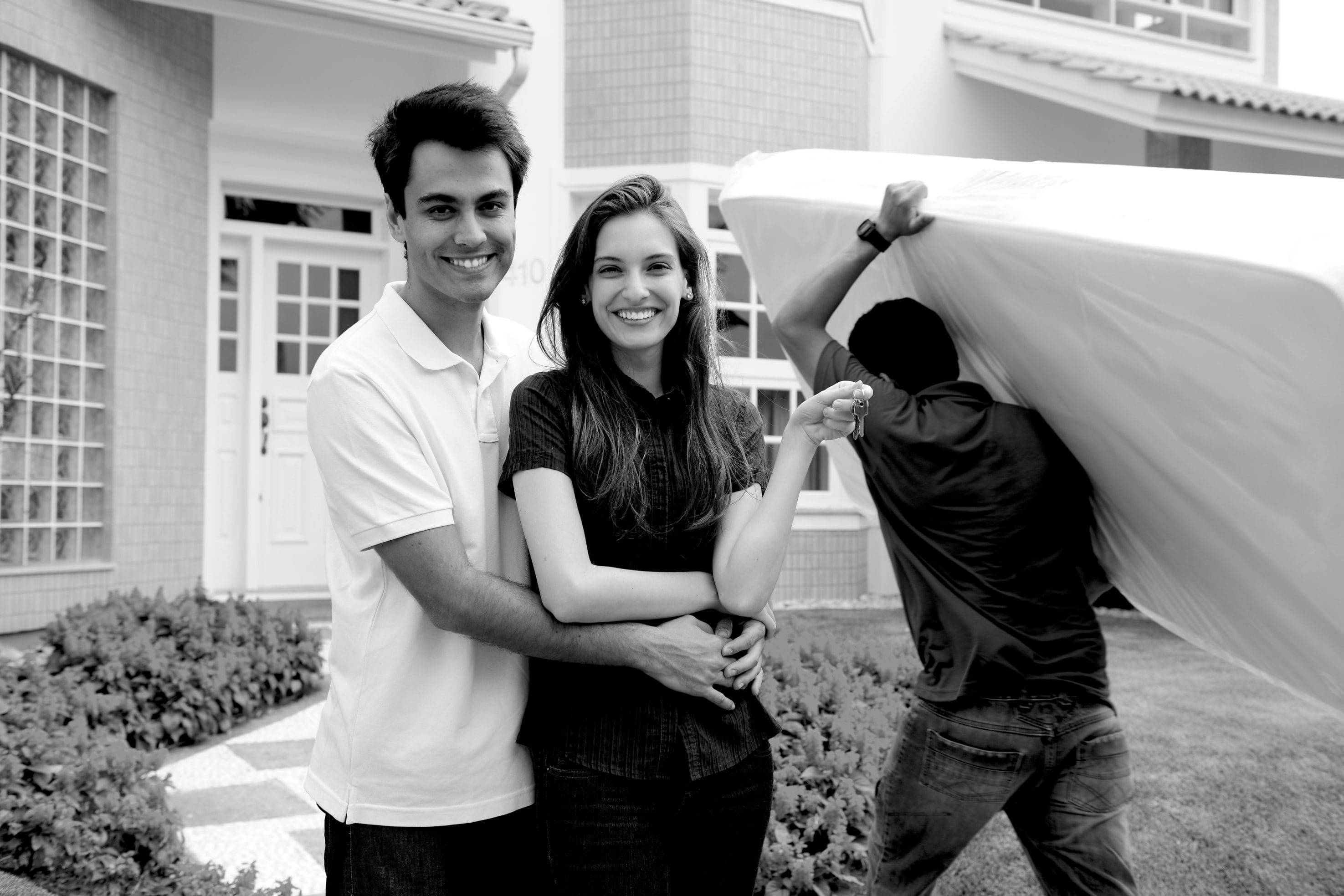 Couple Smiling in Front of New Home with Mover Carrying Mattress
